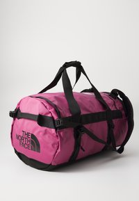 Pink duffel bag with black accents, made of durable fabric, featuring two black handles, adjustable straps, and a logo on the side.