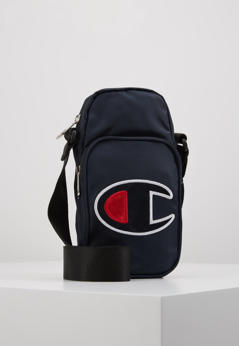 Granatowa torebka na ramię z tkaniny z przednią kieszenią, na której znajduje się duży haftowany logo w białym, czerwonym i czarnym kolorze. W zestawie regulowany czarny pasek.