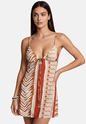 Donna che indossa un vestito senza maniche con motivi a righe verticali nei colori ruggine, beige e bianco, caratterizzato da forme geometriche e da un dettaglio con fiocco davanti.