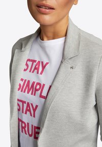 Vrouw wearing een witte t-shirt met rode tekst "STAY SIMPLE STAY TRUE" onder een lichtgrijze blazer met een bijenspeld op de revers.