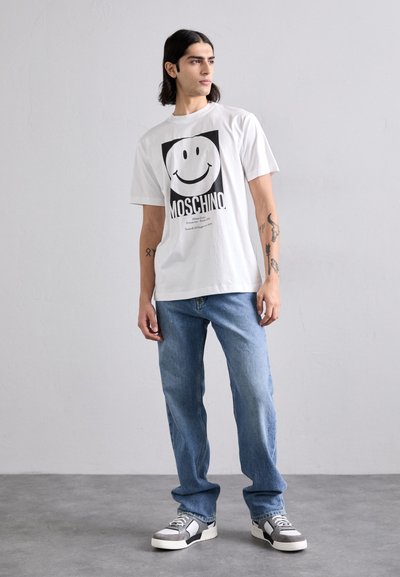 Hvit bomull T-skjorte med en svart smiley-face grafikk og "MOSCHINO" tekst, parret med lyseblå denimjeans og grå joggesko.