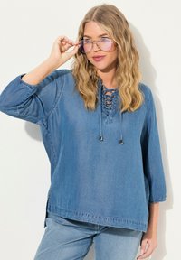 Tunic in denim azzurro chiaro, con scollatura a lacci, maniche a tre quarti e spacchi sui fianchi. Il tessuto ha una finitura morbida e testurizzata.