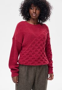Czerwony sweter z dzianiny z fakturowanym wzorem, z okrągłym dekoltem i długimi rękawami. Tkanina wydaje się miękka i lekko puszysta.