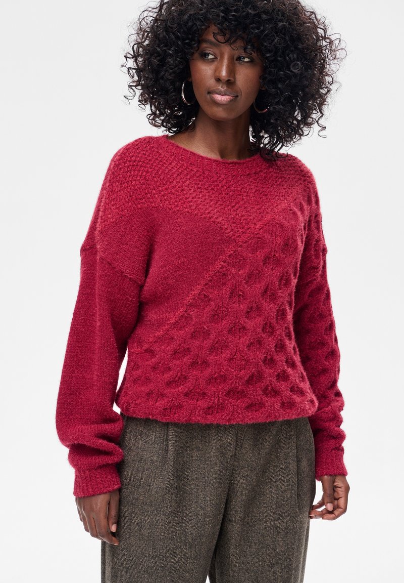 Czerwony sweter z dzianiny z fakturowanym wzorem, z okrągłym dekoltem i długimi rękawami. Tkanina wydaje się miękka i lekko puszysta.
