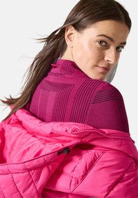 Rosa gepolsterte Jacke mit einem gesteppten Textur, getragen über einem figurbetonten lila Oberteil mit schwarzen Linienmustern und einem hohen Kragen.