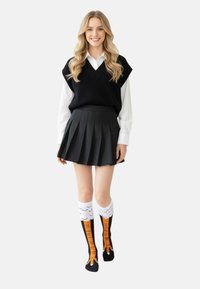 Młoda kobieta z długimi blond włosami nosi czarny sweter bez rękawów, białą koszulę, plisowaną czarną spódnicę oraz skarpety do kolan z designem pomarańczowych pazurów ptaka.