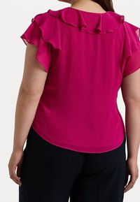 Fuchsias topp med volangärmar och rundad halsringning, tillverkad av lätt tyg; slät yta och lätt glans synlig.