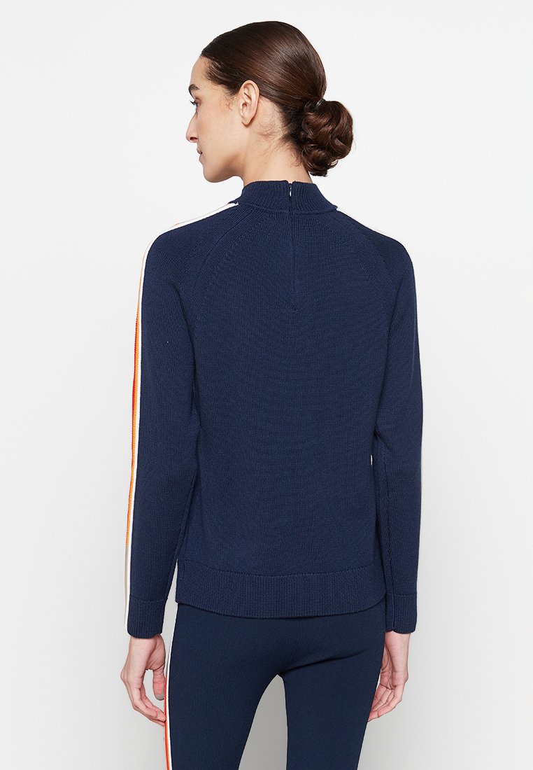 Vrouw met bruin haar in een lage knot, gekleed in een marineblauwe trui met zijstrepen en bijpassende marineblauwe broek, met haar rug naar de camera gekeerd tegen een witte achtergrond.