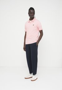 Άνδρας που φοράει ροζ και λευκή ριγωτή μπλούζα πόλο, μπλε παντελόνι, λευκά αθλητικά παπούτσια, στέκεται με το ένα χέρι στην τσέπη μπροστά σε λευκό φόντο.