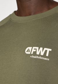 Olive zaļš kokvilnas kreklveida džemperis ar baltu logo "FWT by PeakPerformance" uz krūtīm, ar ribinātu apkakli.