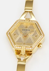 Złoty sześciokątny zegarek z wzorzystą tarczą, ozdobiony cyrkoniami wokół bezel i bransolety, prezentujący teksturowaną powierzchnię oraz elegancki design.