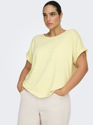 Žena v krátkém žlutobílém pruhovaném tričku a světlých béžových kalhotách, s rukama v kapsách, s tetováním sklenice na martini na pravé paži.