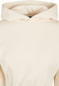 Sweat à capuche de couleur crème avec un design simple, incluant une capuche pliée visible et une texture de tissu douce.