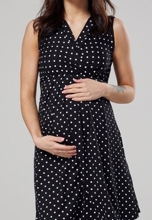 Těhotná žena v černých šatech bez rukávů s bílými puntíky jemně drží břicho oběma rukama na jednobarevném šedém pozadí.