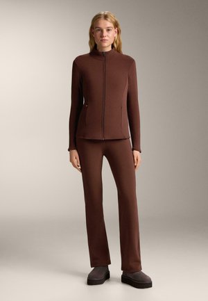 Hnedá bunda so zipsom a vysokým golierom a zodpovedajúce priliehavé nohavice. Hladká, elastická tkanina. Model nosí sivé platformové topánky.