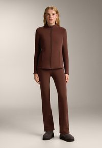 Smeđa jakna na raszip s visokim okovratnikom i odgovarajuće prilagođene hlače. Glatka, rastezljiva tkanina. Model nosi sive platforme.