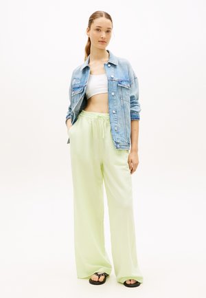 Jeune femme debout portant une veste en jean bleue par-dessus un crop top blanc, un pantalon large vert clair et des sandales noires.