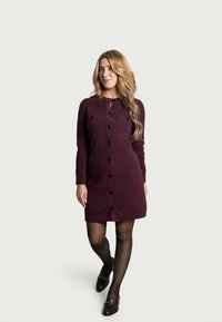 Strickkleid mit langen Ärmeln in tiefem Burgunderrot, Knopfverschluss und zwei Brusttaschen. Mit glatter Textur und knielanger Schnittform.