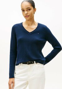 Suéter de punto azul marino con escote en V y mangas largas, de textura suave, con un pequeño logo en el pecho, combinado con pantalones blancos y un cinturón negro.
