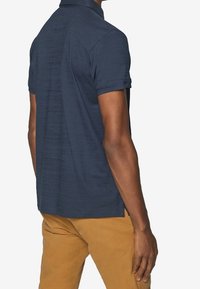 Polo shirt bleu marine à manches courtes avec un tissu texturé, un col doux et des poignets repliés, porté avec un pantalon beige clair, montrant la vue arrière.