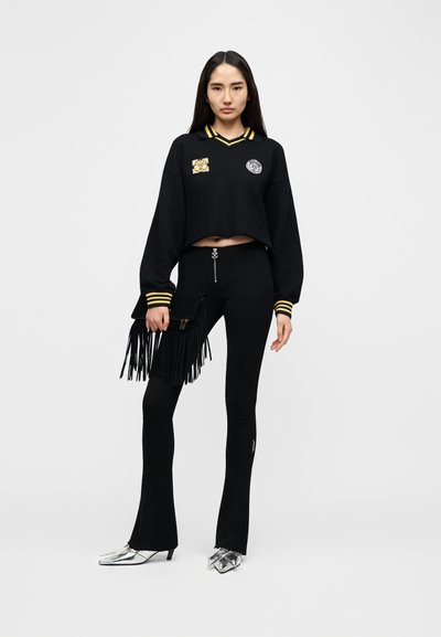 Μαύρο κοντό φούτερ με κίτρινο ριγωτό γιακά και μπαλώματα, συνδυασμένο με μαύρο καμπάνα παντελόνι, ασημένια μυτερά παπούτσια και μαύρο φάκελο με φράντζες.