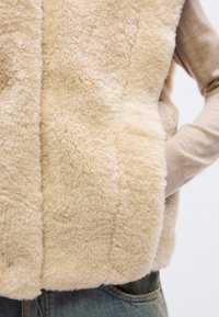 Μπεζ γιλέκο από ψεύτικη γούνα με απαλή, αφράτ η υφή, με ψηλό γιακά και πλαϊνές τσέπες, που φοριέται πάνω από ένα ανοιχτό μπεζ μακρυμάνικο τοπ.