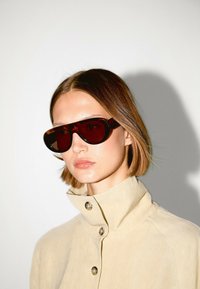 Okulary przeciwsłoneczne o brązowym odcieniu z zaokrąglonym kształtem, noszone z jasnobeżową kurtką zapinaną na guziki z kołnierzykiem. Gładka faktura i minimalistyczny design.