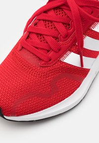 Botte de sport rouge avec une tige en maille texturée, des lacets rouges et des accents blancs, dotée d'une semelle extérieure noire et d'un embout en caoutchouc lisse.