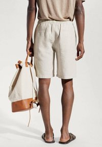 Shorts en lin beige avec une ceinture élastique, accompagnés d'un sac à dos beige et marron; des sandales complètent ce look décontracté.