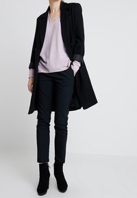 Cappotto lungo nero, maglione leggermente viola a scollo a V, pantaloni scuri e stivali neri alla caviglia. Il cappotto ha le maniche arrotolate; gli stivali presentano un dettaglio con zip.