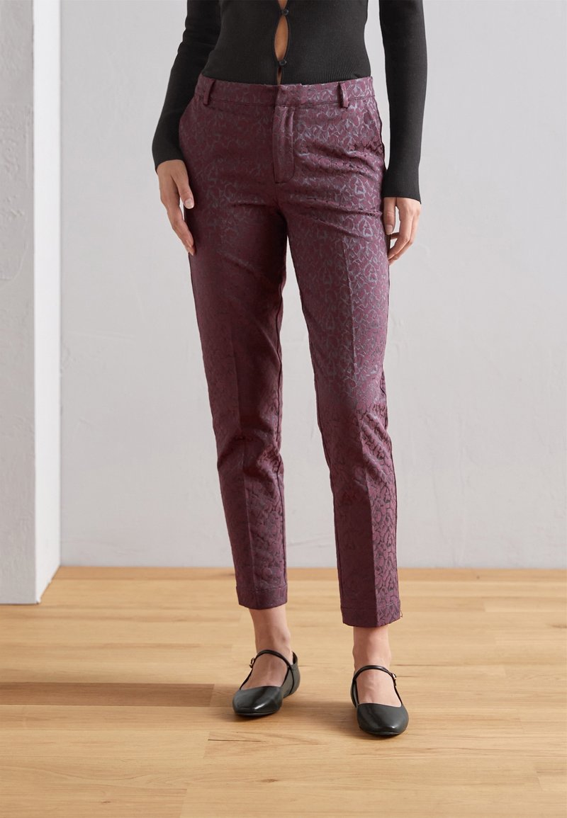 Femme portant un pantalon ajusté violet foncé à motifs longueur cheville avec des ballerines Mary Jane noires sur un sol en bois.