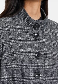 Cappotto a quadri con colletto alto, tessuto grigio e nero strutturato, e quattro bottoni neri sul davanti.