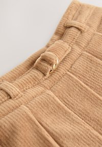 Tissu en velours côtelé couleur fauve avec nervures verticales, orné d'un anneau décoratif doré et d'une lanière. Texture douce, vue en gros plan.