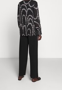 Blusa nera a maniche lunghe con motivo floreale bianco, vestibilità ampia e schiena plissettata. Abbinata a pantaloni neri a gamba larga e sandali piatti.