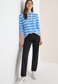 Sweat à capuche rayé bleu et blanc avec une poche avant, pantalon noir à jambes droites, et baskets blanches avec des accents de couleur. Tissu lisse et décontracté.