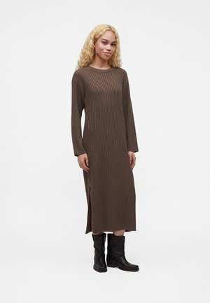 Femme aux cheveux blonds bouclés portant une longue robe marron côtelée avec une fente latérale et des bottes noires mi-mollet, debout sur fond blanc.