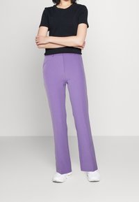 Person som står med armarna i kors, iförd en svart kortärmad skjorta, lila rakbenade byxor och vita sneakers på en enfärgad bakgrund.