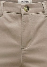 Pantalon beige avec fermeture à boutons, passants pour ceinture et étiquette de la marque Only & Sons visible sur la taille.