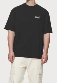 Czarny bawełniany t-shirt z krótkim rękawem, okrągłym dekoltem i małym białym logo na piersi. Materiał wydaje się gładki i lekki.