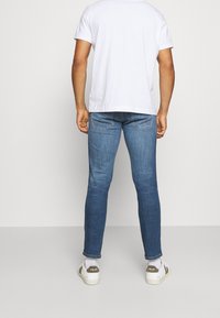 Blue slim-fit jeans with a faded finish, five-pocket design, and stitching details, paired with a plain white t-shirt and white sneakers.