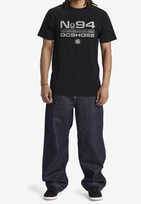 Svart bomull t-shirt med silvertryck "No 94 DC Shoes" på bröstet, kombinerad med löst sittande mörk denimjeans och svarta sneakers.
