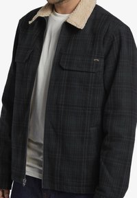 Black and green plaid jacket with a beige sherpa collar, zippered front, and two chest pockets. Textured fabric with a relaxed fit.