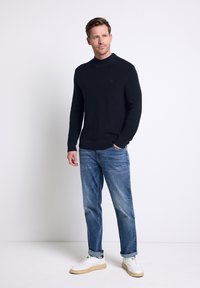 Pull en maille noir avec un col haut, associé à un jean bleu clair et des baskets blanches avec des accents verts. Design simple et décontracté.
