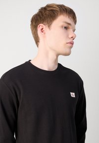 Young man with short light brown hair wearing a black long-sleeve shirt with a small Calvin Klein logo on the chest.