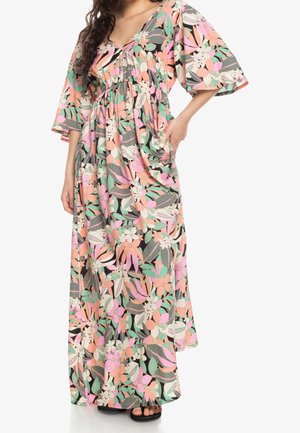 Femme portant une longue robe fleurie avec des feuilles et des fleurs roses, pêche, vertes et blanches, avec des manches 3/4 et un décolleté en V.