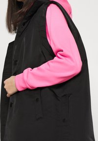 Svart vadderad väst med knappar, lagrad över en ljusrosa sweatshirt. Vesten har en slät textur och en avslappnad passform.