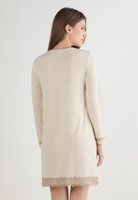 Frau mit braunen Haaren, die ein beiges Langarmkleid mit Spitzenbesatz an den Ärmeln und dem Saum trägt, vor einem schlichten weißen Hintergrund stehend.