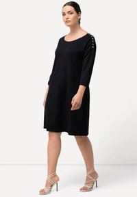 Abito blu navy a lunghezza ginocchio con maniche a tre quarti, dotato di bottoni sulle spalle, tasche laterali e una texture di tessuto fluido e drappeggiato.