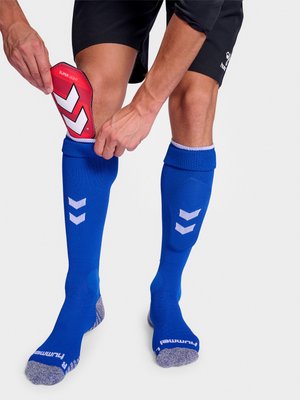 Person wearing blue sports socks inserts a red "SUPER LIGHT" shin guard into the left sock, wearing black shorts on a white background.