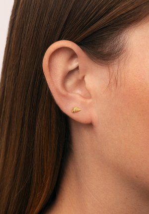Pendiente de oro en forma cónica con acentos texturizados, llevado en el lóbulo de la oreja de un modelo, que presenta piel suave y cabello largo.